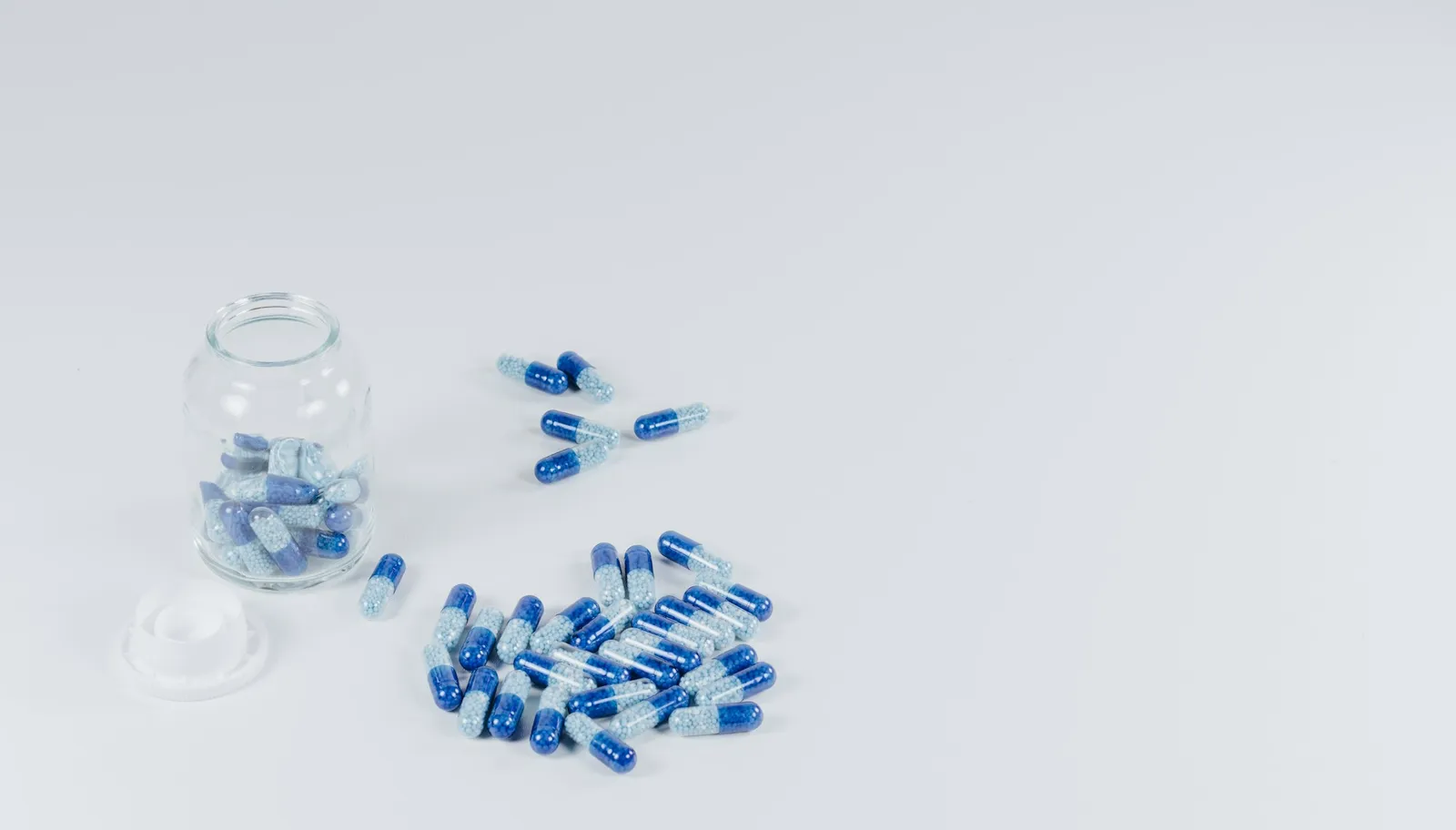 Blue and light blue supplement capsules beside a glass jar of capsules on a white table.