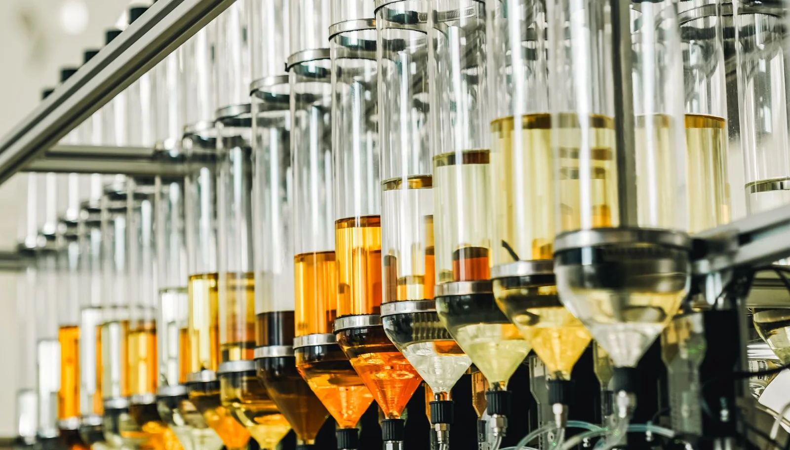 Rows of clear dispensing cylinders containing yellow and amber liquids on automated bottling machinery.