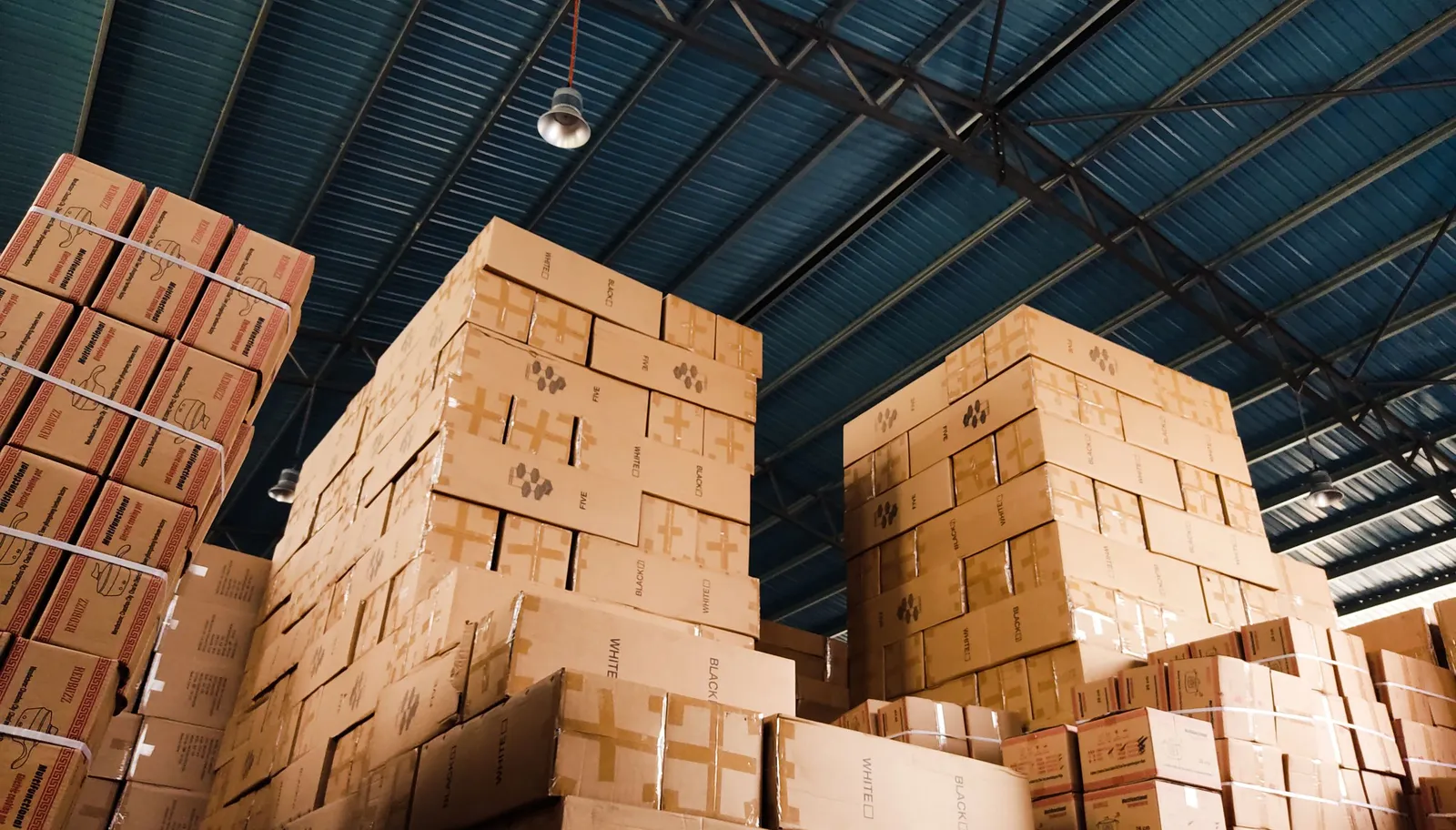 High stacks of shipping cartons in a warehouse, each needing case-level barcode labels